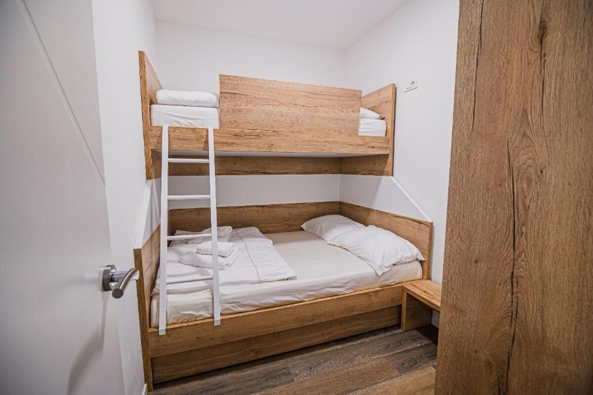 Uma cama beliche com estrutura de madeira e roupa de cama branca, uma escada e um pequeno banco de madeira.