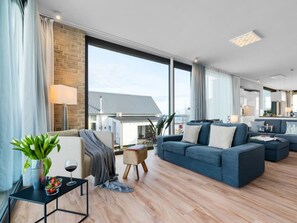 Ein modernes Wohnzimmer mit einer Couch, einem kleinen Tisch mit Pflanze und einem Weinglas, und Blick auf ein Haus durch große Fenster.