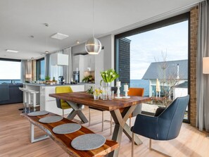 Cozinha moderna com ilha central, área de jantar com mesa e bancos de madeira, e uma grande janela com vista para o mar.