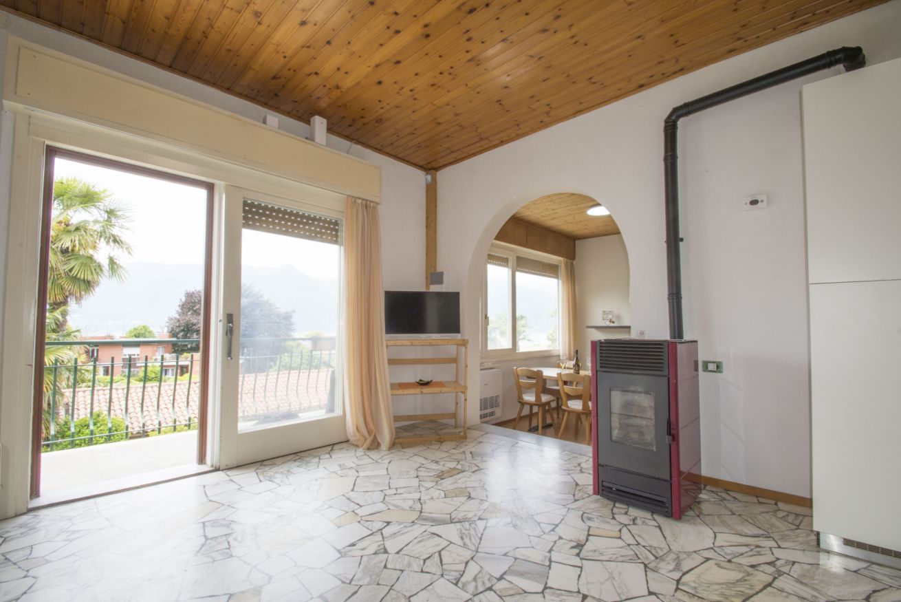 A spacious room with a stone-patterned floor, a wooden ceiling, a television, a fireplace, and a balcony with a view of greenery.