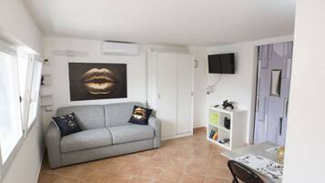 Una sala de estar con un sofá gris, un televisor montado en la pared, una estantería y una puerta.