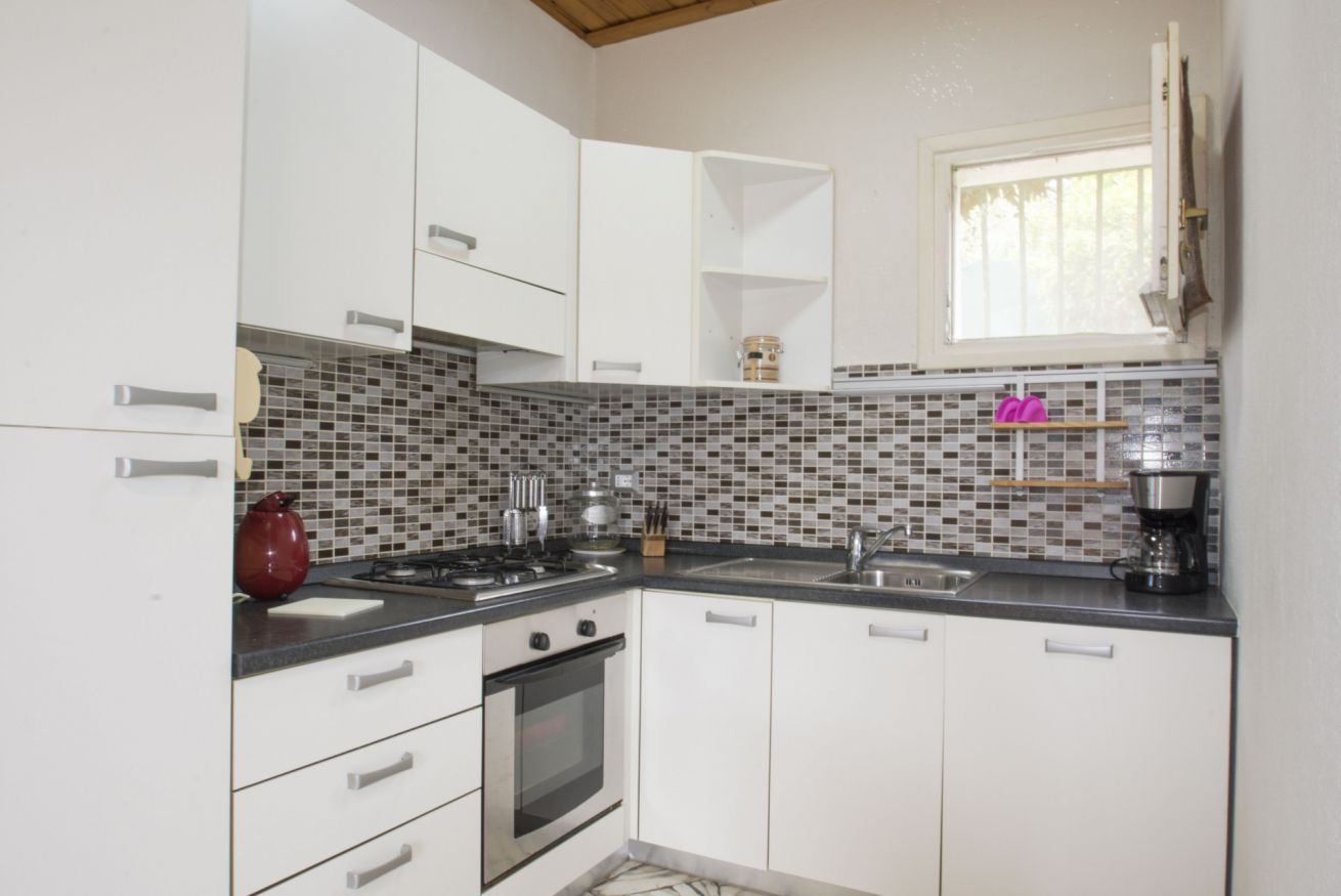 Una cucina compatta con mobili bianchi, zoccolo in piastrelle, piano cottura a gas e macchina per il caffè.