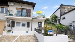 A two-story residential building with a balcony, a paved driveway, and a small garden area.