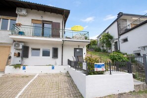 A two-story residential building with a balcony, a paved driveway, and a small garden area.