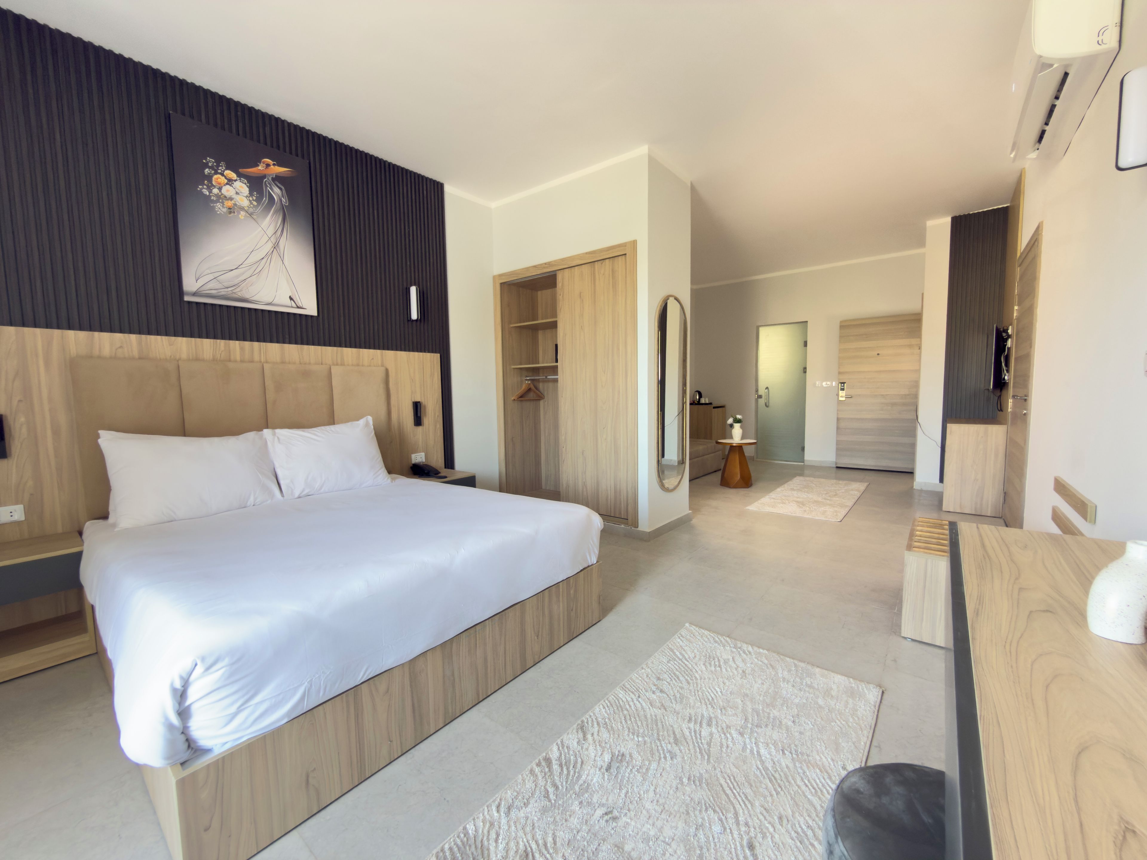 Ett modernt hotellrum med en stor säng, ett träfärgat sänggavel, ett nattduksbord, en spegel och ett litet runt bord med en vas.
