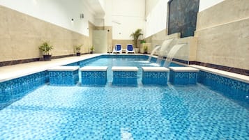 An indoor swimming pool with a tiled floor, a water feature, and lounge chairs.
