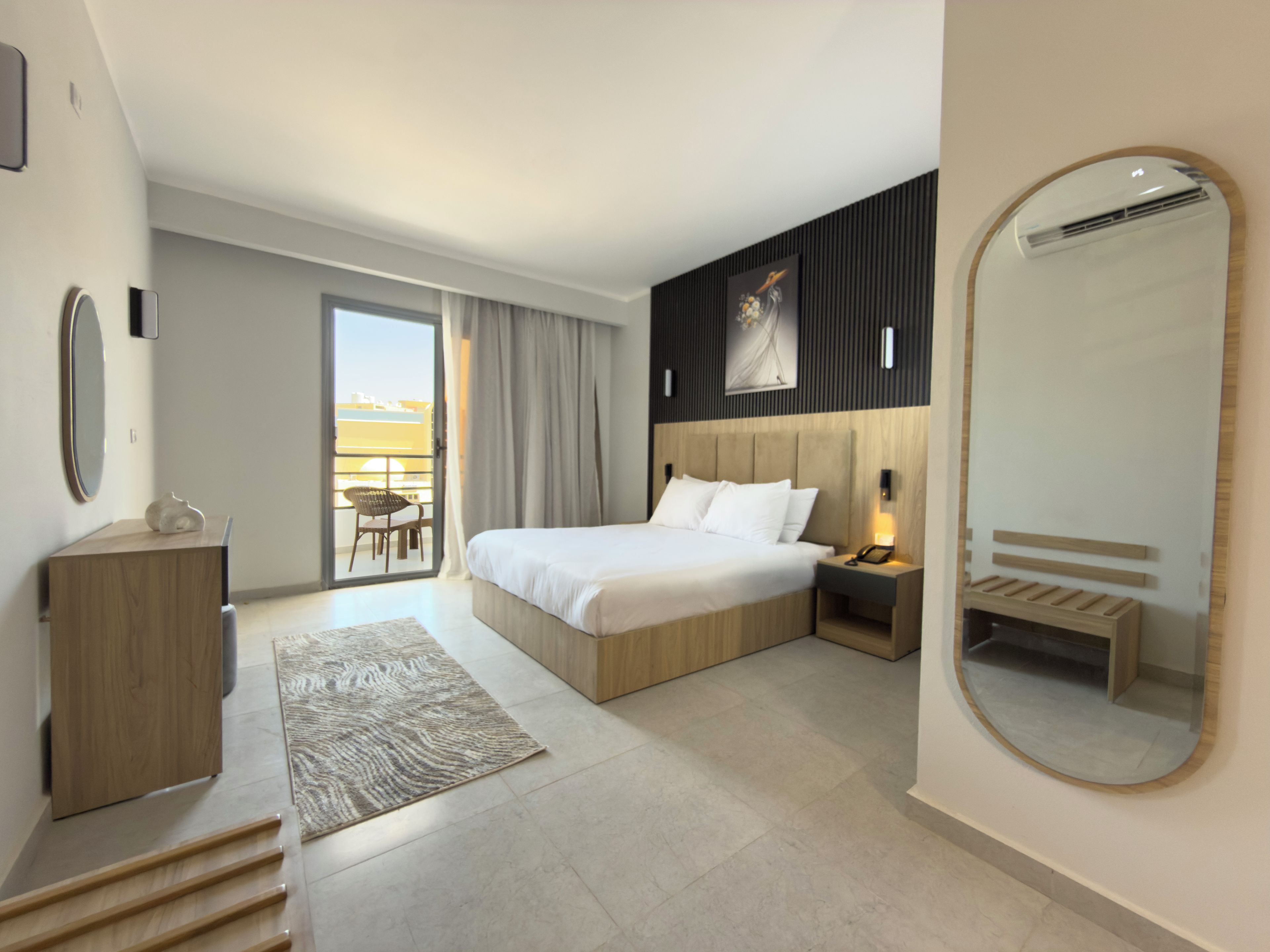 A modern hotel room with a large bed, a bedside table with a lamp, a round mirror, a wooden bench, and a view of the outdoors.