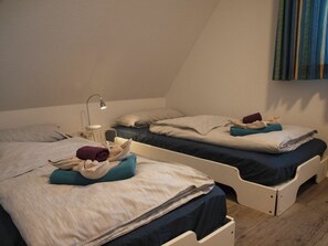 Ein Zimmer mit zwei Betten, jeweils mit Kissen und Decke. Ein Nachttisch mit Lampe und einem kleinen Tisch sind vorhanden.