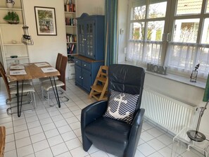 Ein Zimmer mit einem Esstisch, Stühlen, einem schwarzen Ledersofa mit einem gemusterten Kissen, einem blauen Schrank und einem Fenster mit Vorhängen.
