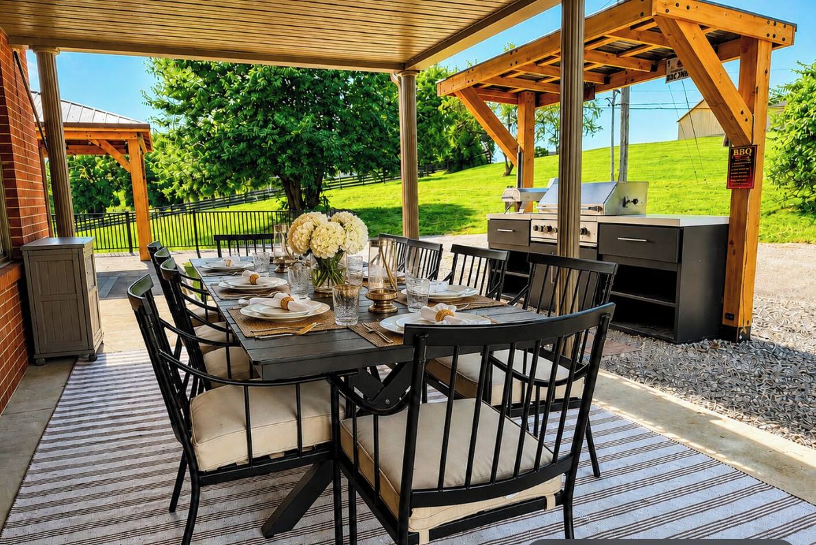 An outdoor dining area with a covered table and chairs, a grill, and a view of a green landscape.