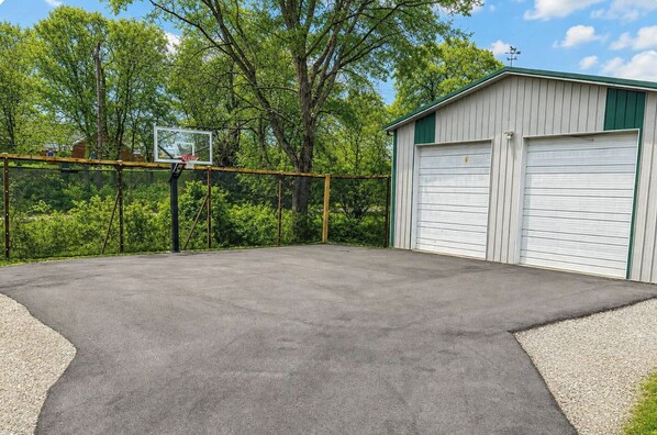 A basketball court with a hoop, surrounded by trees and a garage with two doors.