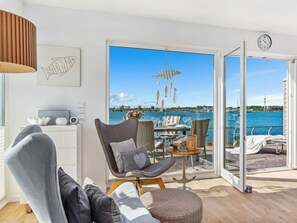 Ein modernes Wohnzimmer mit Blick auf ein Gewässer, ein Essbereich mit Tisch für Zwei und eine gemütliche Sitzecke mit Sessel und Hocker.
