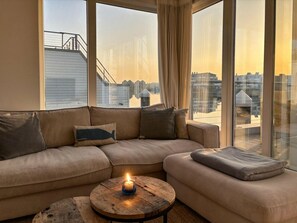A modern living room with a sectional sofa, a round coffee table with a candle, and large windows offering a city view. - Houseboat in Olpenitz With Swimming Pool (Kappeln)