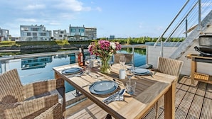 A dining area with a table set for two, overlooking a body of water and modern buildings in the background. - Houseboat in Olpenitz With Swimming Pool (Kappeln)