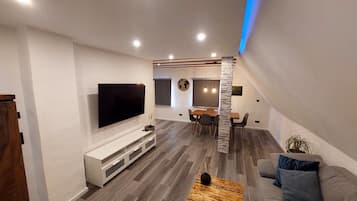 Ein modernes Wohnzimmer mit einem Flachbildfernseher, Holzböden, einem Essbereich mit Tisch und Stühlen sowie einer Treppe, die in eine obere Ebene führt.