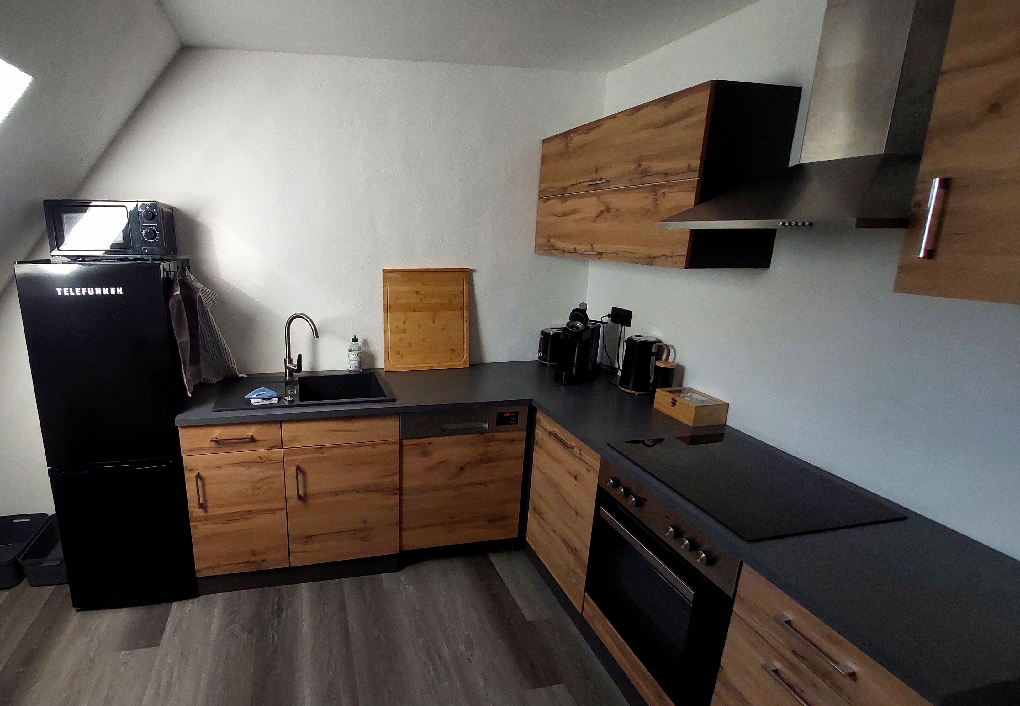 Eine kleine Küche mit einem schwarzen Kühlschrank, Holzschränken, einem Spülbecken und einem Herd.