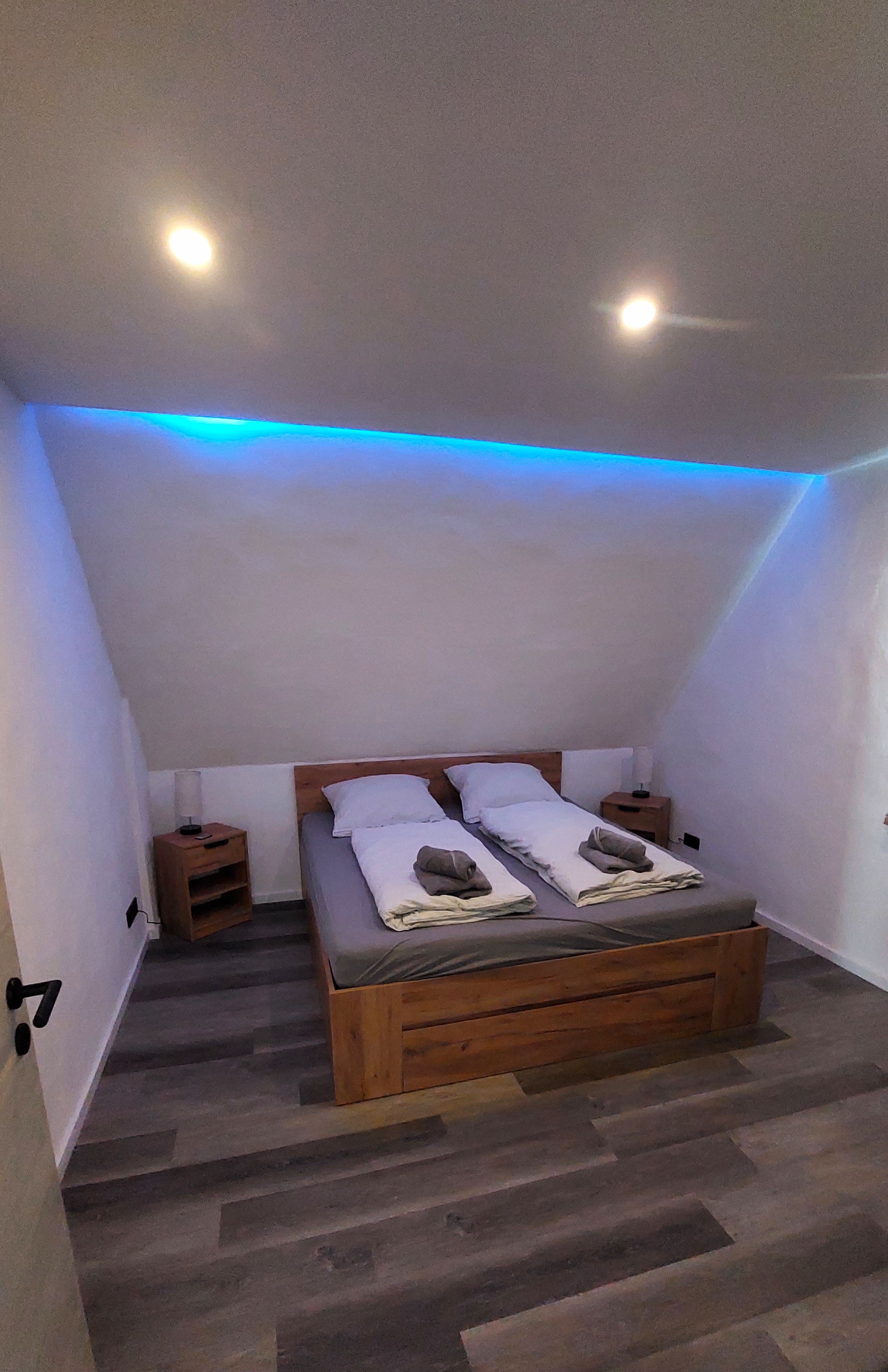 Ein Schlafzimmer mit einem Holzbett, zwei Nachttischen und einer schrägen Decke mit blauer Beleuchtung.