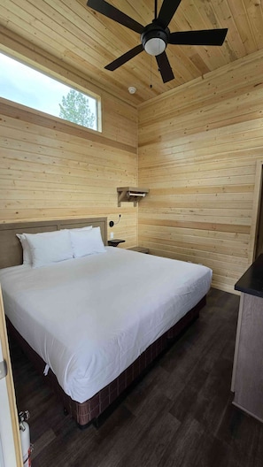 Ein Schlafzimmer mit Holzwänden, einem Bett, einem Nachttisch, einem Deckenventilator und einem Fenster.