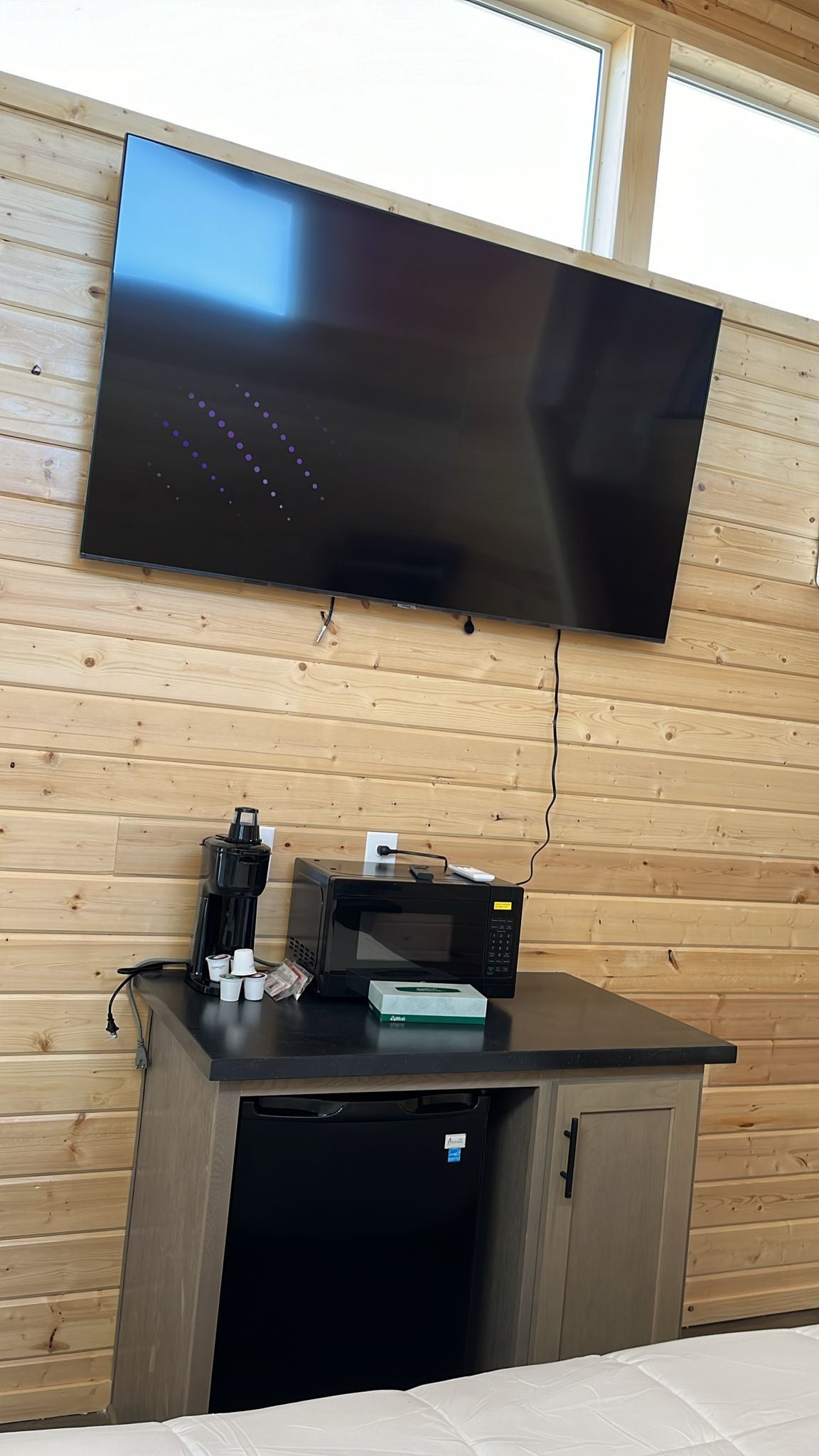 A modern room with a flat-screen TV mounted on a wooden wall, a coffee maker, a microwave, and a small refrigerator.