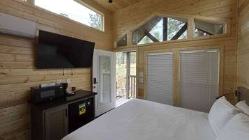 Un dormitorio de cabaña de madera con una cama, un televisor montado en la pared, un escritorio pequeño con cafetera y una ventana con vistas a un bosque.