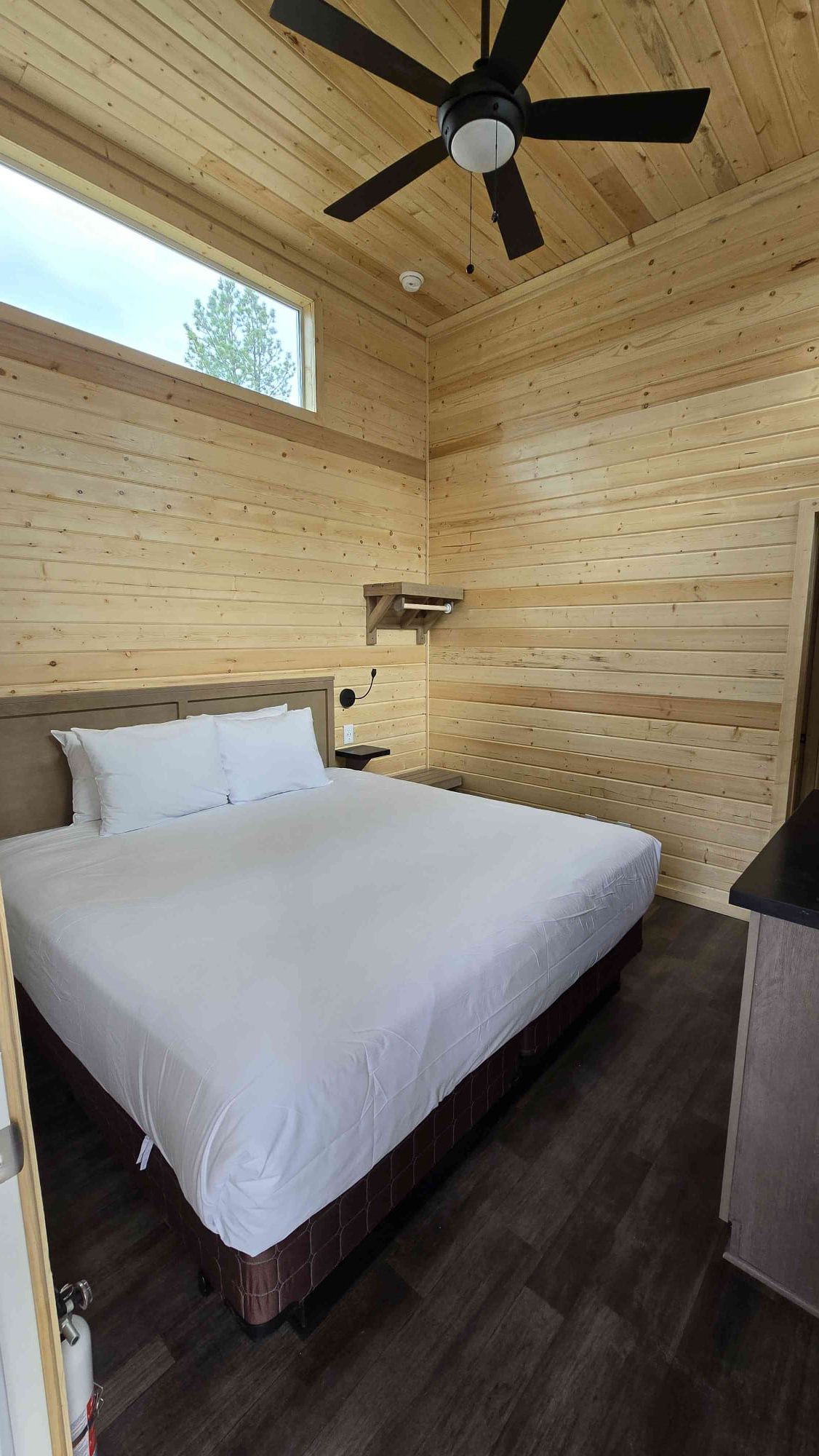 Un dormitorio con paneles de madera, una cama, una mesita de noche, un ventilador de techo y una ventana.