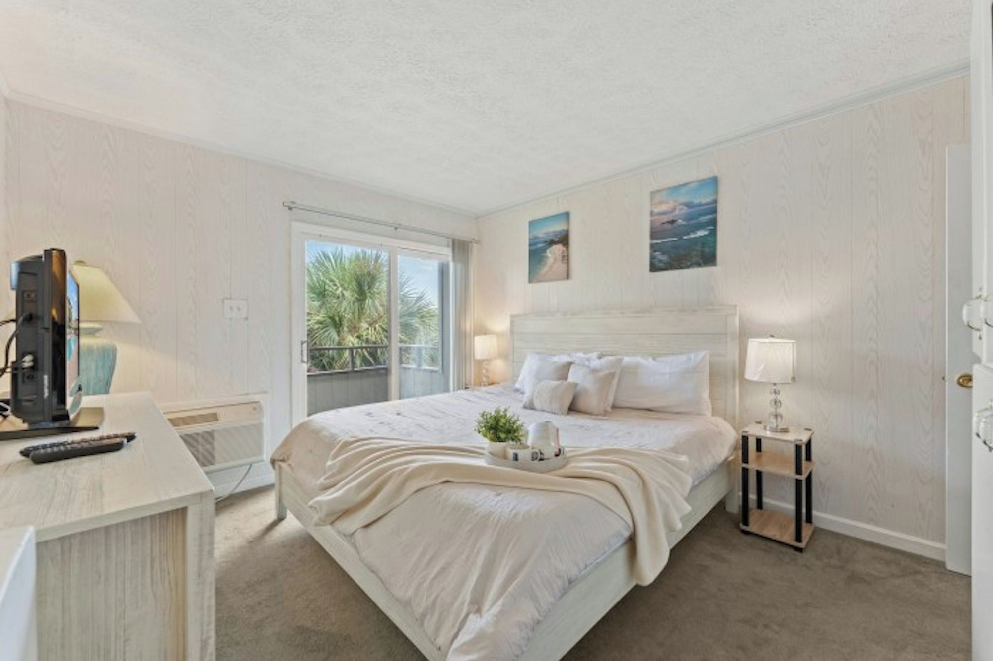 A hotel room with a bed, bedside table, lamp, and a view of the outdoors.