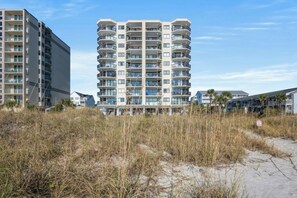 A multi-story residential building with balconies, situated near a sandy area with sparse vegetation. - Crescent Towers II 508-Oceanfront Corner Unit-2BR (North Myrtle Beach)