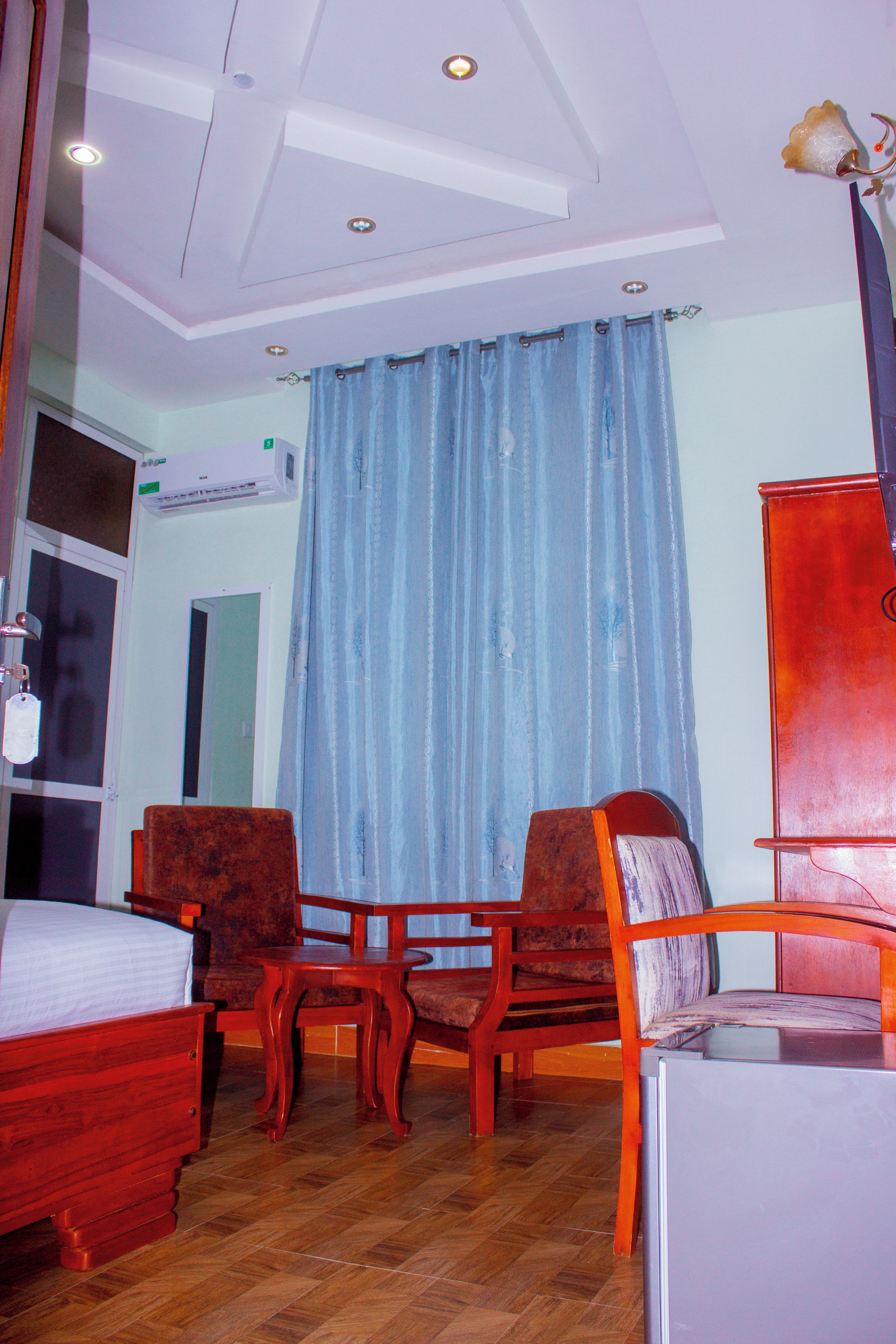 Ein Hotelzimmer mit einem Bett, zwei Holzstühlen, einem kleinen Tisch, einem Schrank und einem Fenster mit Vorhängen.