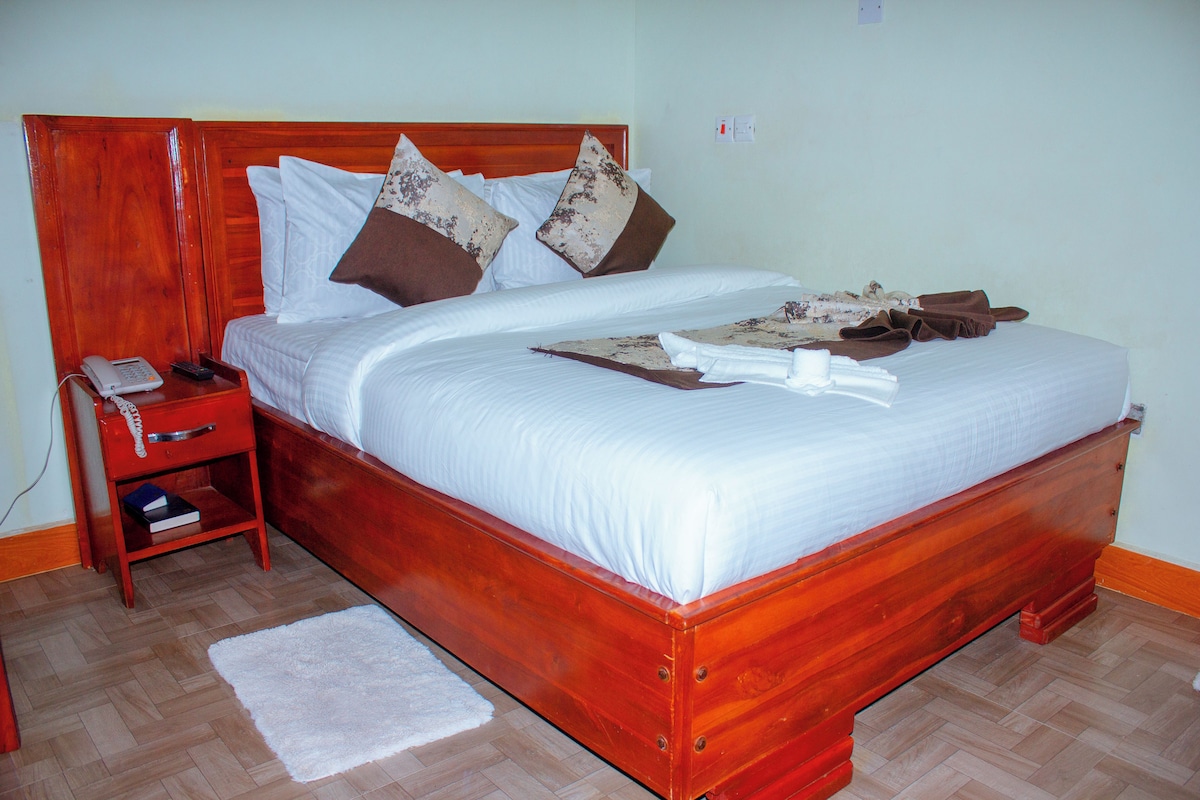 Cama bem arrumada com cabeceira de madeira, mesa de cabeceira com telefone e um tapete pequeno no chão.