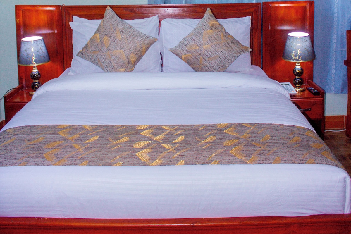 Quarto de hotel padrão com cama de madeira, duas luminárias laterais e colcha estampada.