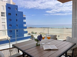 En balkon med havudsigt, et bord med bog og glas, samt en blå bygning i baggrunden.