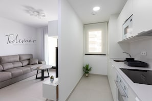 Eine moderne Wohnung mit einem grauen Sofa, einem kleinen weißen Tisch, einer Topfpflanze und einer Küche mit weißen Schränken und einer schwarzen Arbeitsplatte.