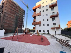 Ein Spielplatz mit Schaukeln und einer Rutsche, umgeben von einem befestigten Bereich mit Bänken.