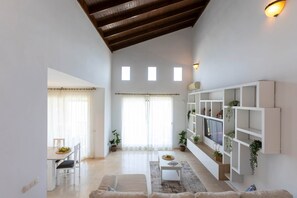 Amplia sala de estar con techo alto, una mesa de comedor con sillas, un sofá, un televisor montado en una pared blanca y un mueble empotrado con plantas.