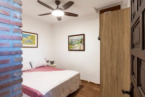 A bedroom with a bed, a ceiling fan, two framed paintings, a brick wall, and a wooden door. - Todimer Luxury Villa Herradura (Almuñécar)