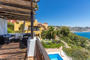 Una terraza con mobiliario exterior, piscina y vistas a un pueblo costero.
