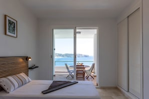 Ein Schlafzimmer mit einem Bett, einem Nachttisch, einem Tischchen, einem gerahmten Bild an der Wand und Meerblick durch eine offene Tür.