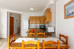 A dining area with a wooden table and chairs, a kitchen with wooden cabinets and appliances, and a small potted plant on the table. - Todimer Neptuno Beach (Torrenueva Costa)