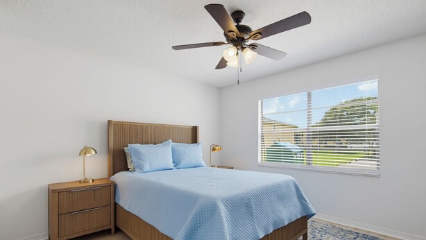 Une chambre à coucher comprenant un lit, des tables de chevet, un ventilateur de plafond, une fenêtre avec des stores et un tapis à motifs.