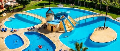Un complejo de piscinas grande con varias secciones y un tobogán de agua, rodeado de tumbonas y sombrillas.