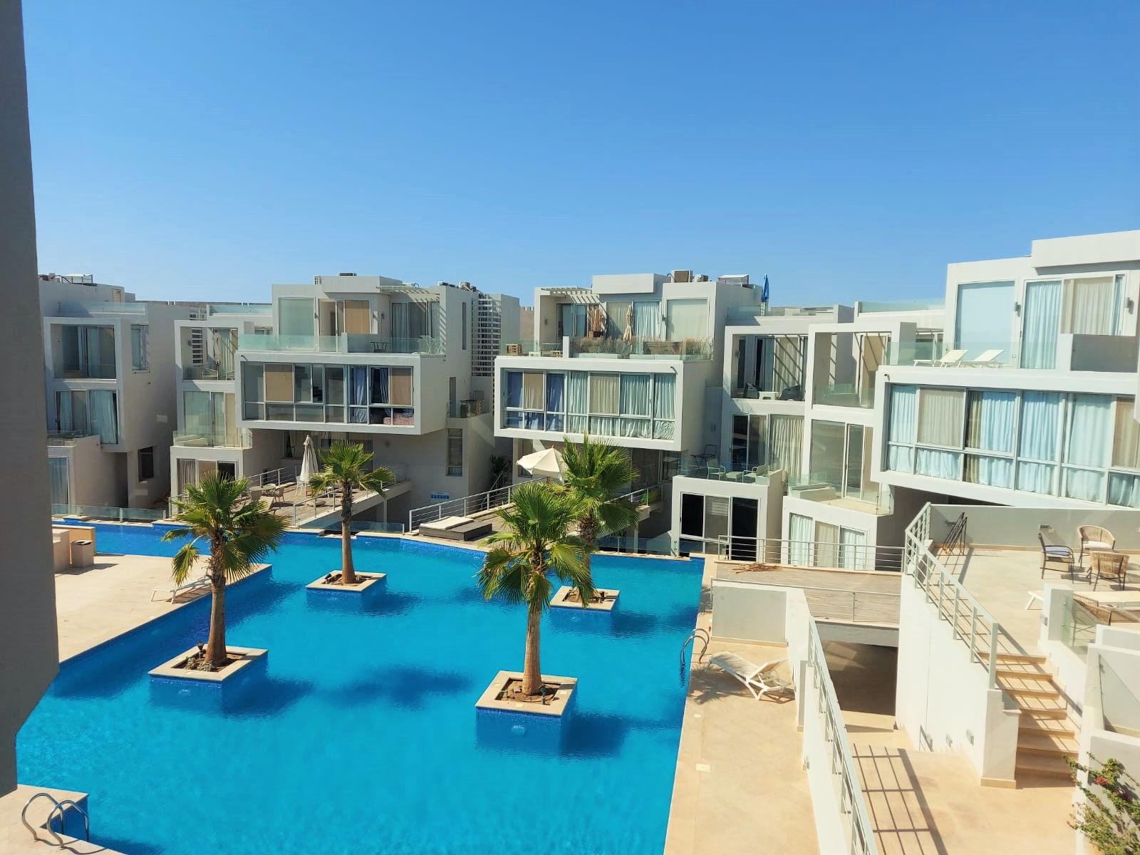 A modern hotel with a central swimming pool surrounded by palm trees and multiple balconies.