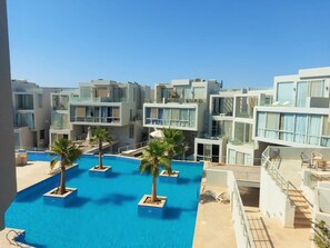 A modern hotel with a central swimming pool surrounded by palm trees and multiple balconies.