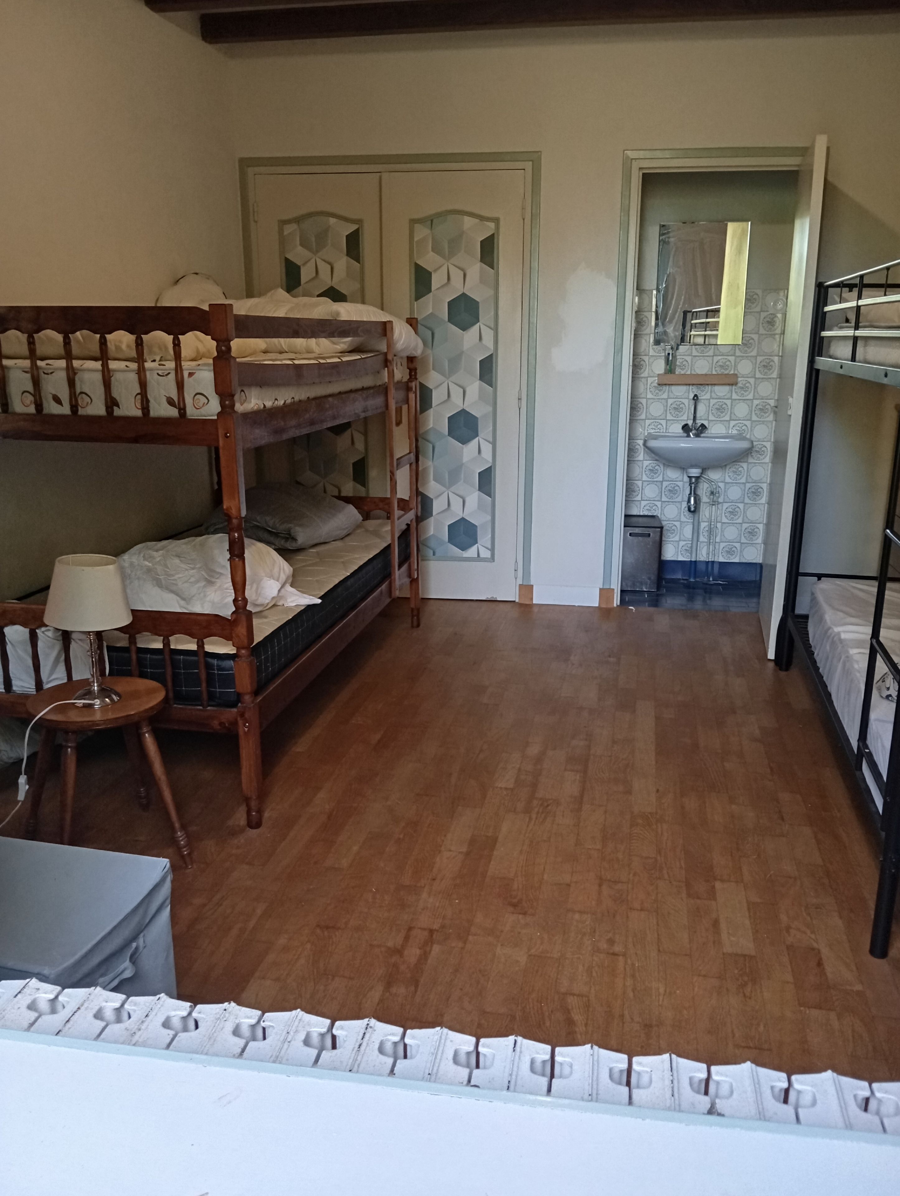 Et værelse med to køjesenge, et lille bord med en lampe, en dør med geometriske mønstre, og et håndvask i det tilstødende rum.