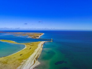 Vista aérea de una zona costera con un faro, aguas azules cristalinas y una playa arenosa.
