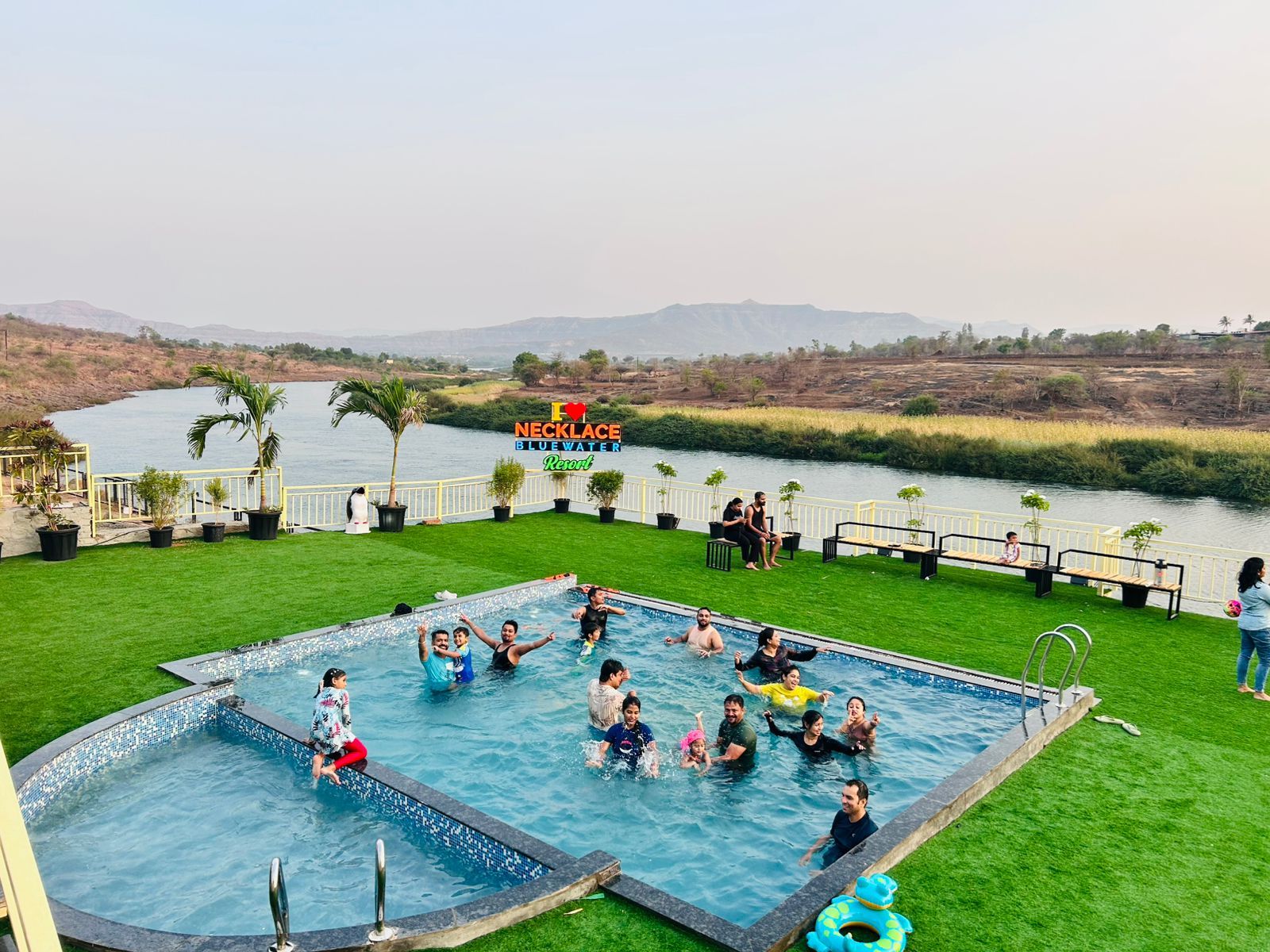 A swimming pool with people enjoying the water, surrounded by a grassy area and a view of a river.