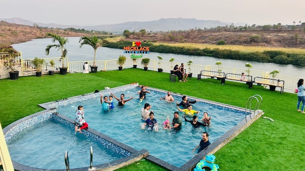 A swimming pool with people enjoying the water, surrounded by a grassy area and a view of a river. - Necklace Bluewater Resort Bhor (Sangamner)