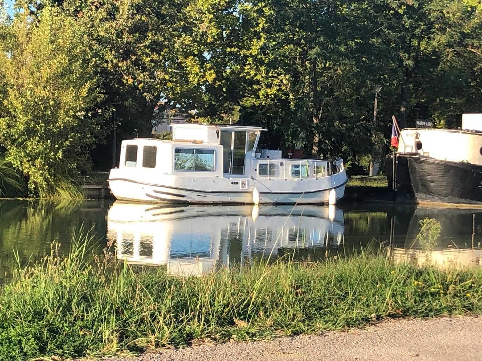 A narrow waterway with a white boat docked on the left, a smaller boat on the right, and dense greenery along the banks.