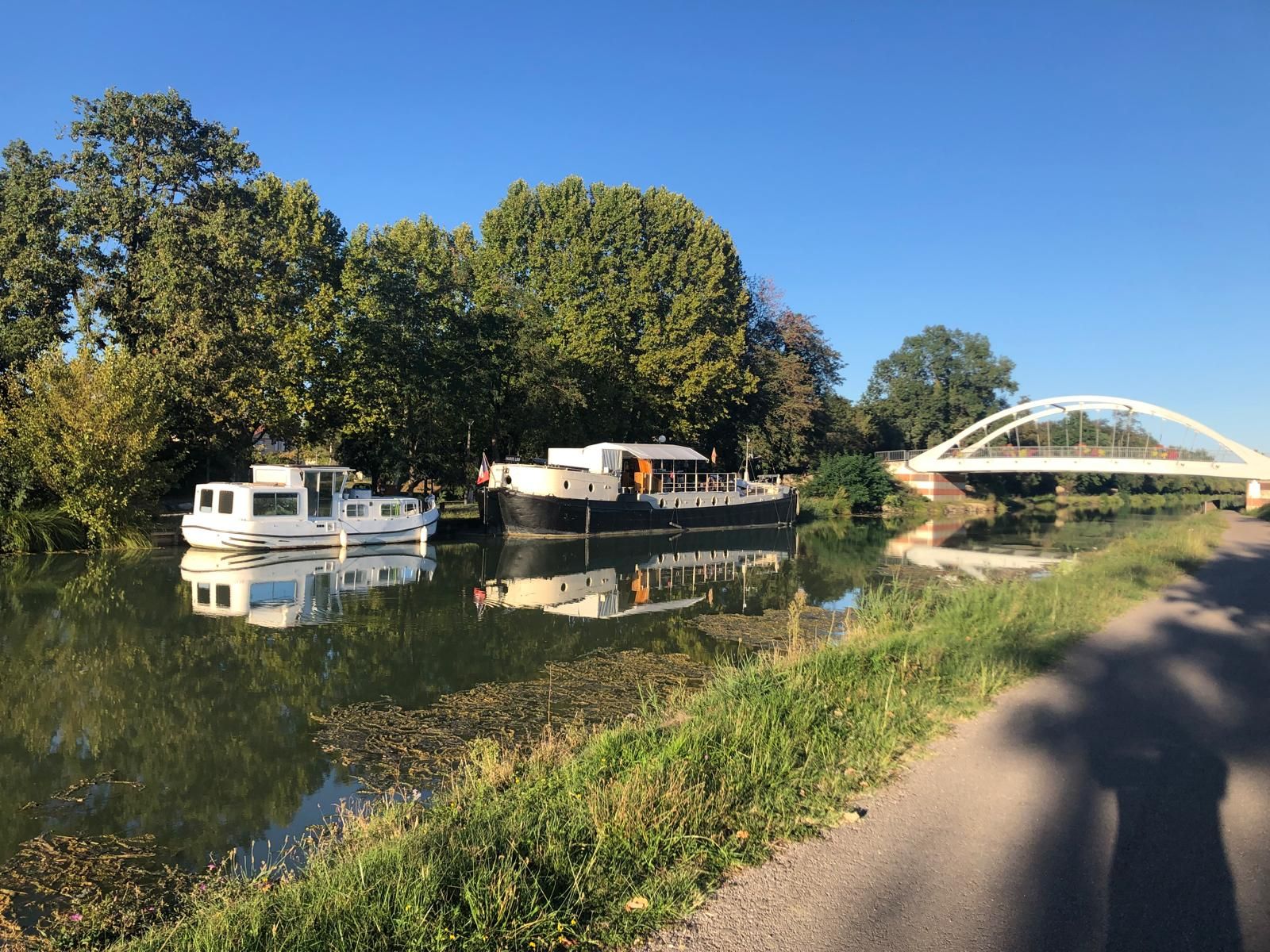 Ein ruhiger Fluss mit angestaubten Booten, ein Fußweg und eine Brücke im Hintergrund.