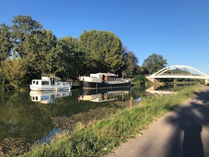 A calm river with boats docked, a pedestrian path, and a bridge in the background.