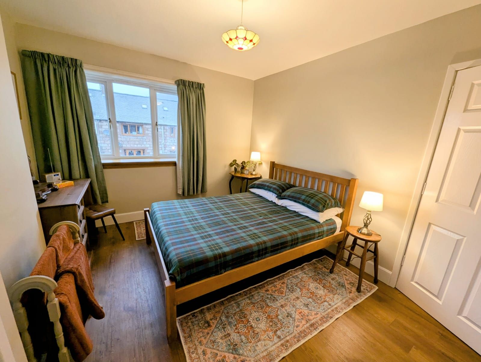 A bedroom with a bed, a desk, a chair, a window with curtains, a lamp, and a door.